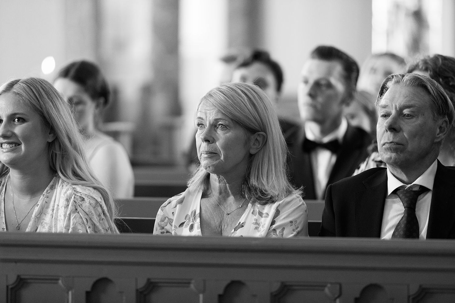 Wedding guests sitting in church pews smile and react during an emotional ceremony moment.