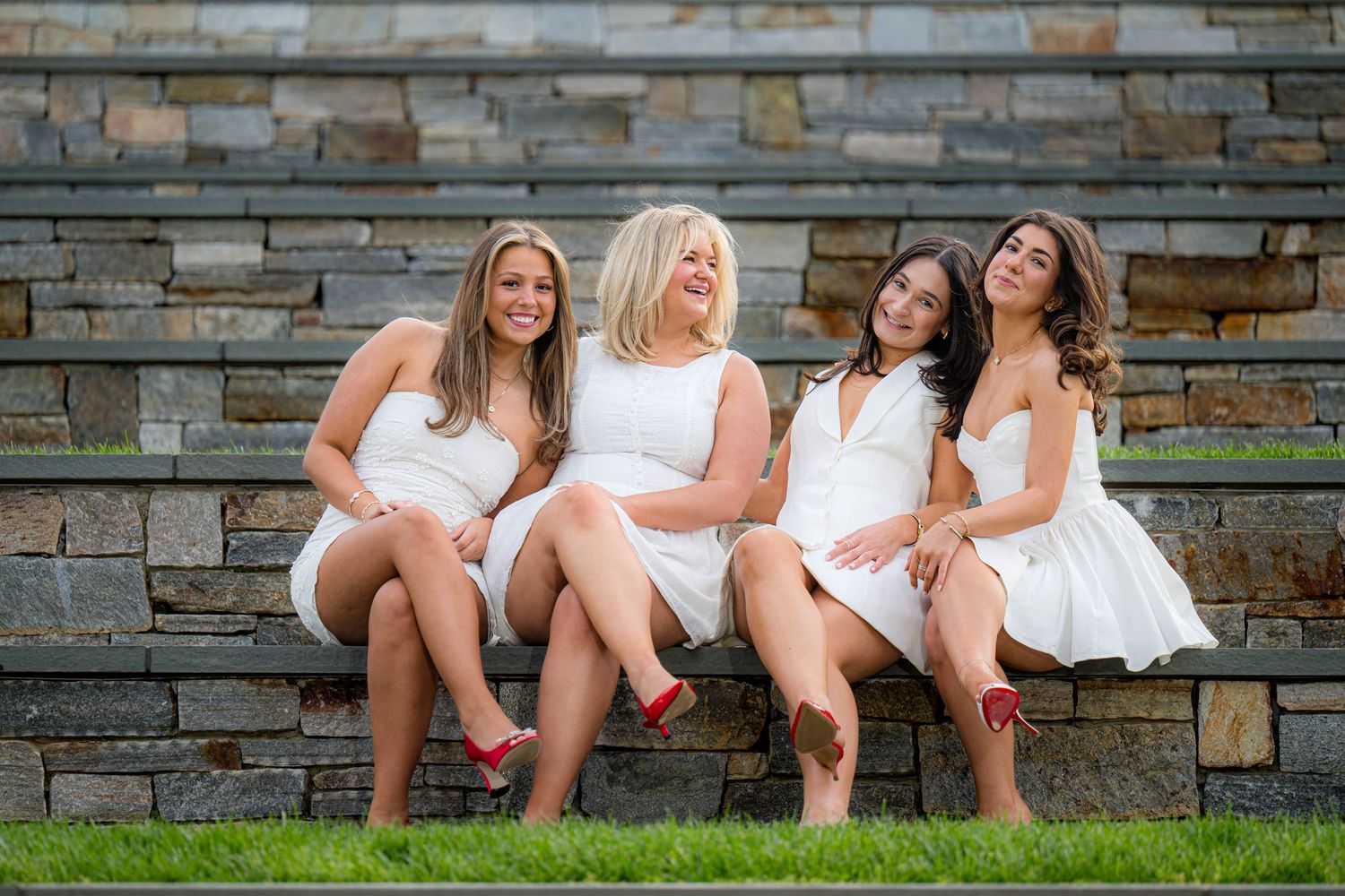 Graduates in white dresses and black gowns throw their caps in celebration under a stone archway.