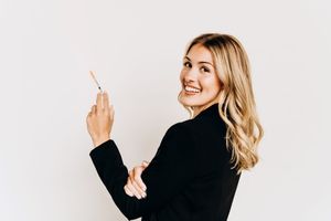 Smiling blonde woman in black blazer poses while holding a small pen or tool, looking back.