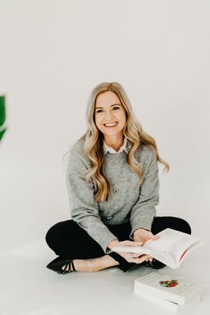 Smiling blonde woman sitting cross-legged on white floor holding an open book, wearing gray sweater and black pants.