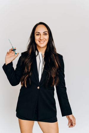 Smiling woman in black blazer holds a medical syringe while posing against a white background.
