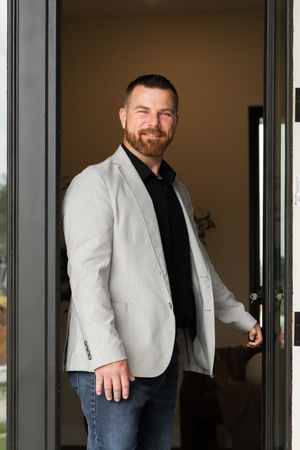 Smiling bearded man in gray blazer and black shirt standing in an open glass doorway.