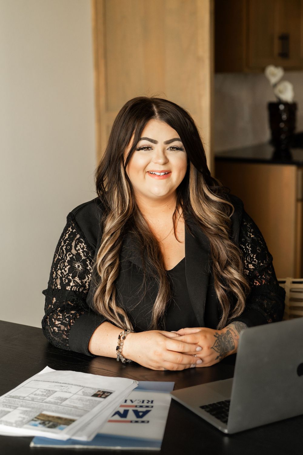 Smiling female real estate agent in black lace top sitting at desk with laptop and property brochures.