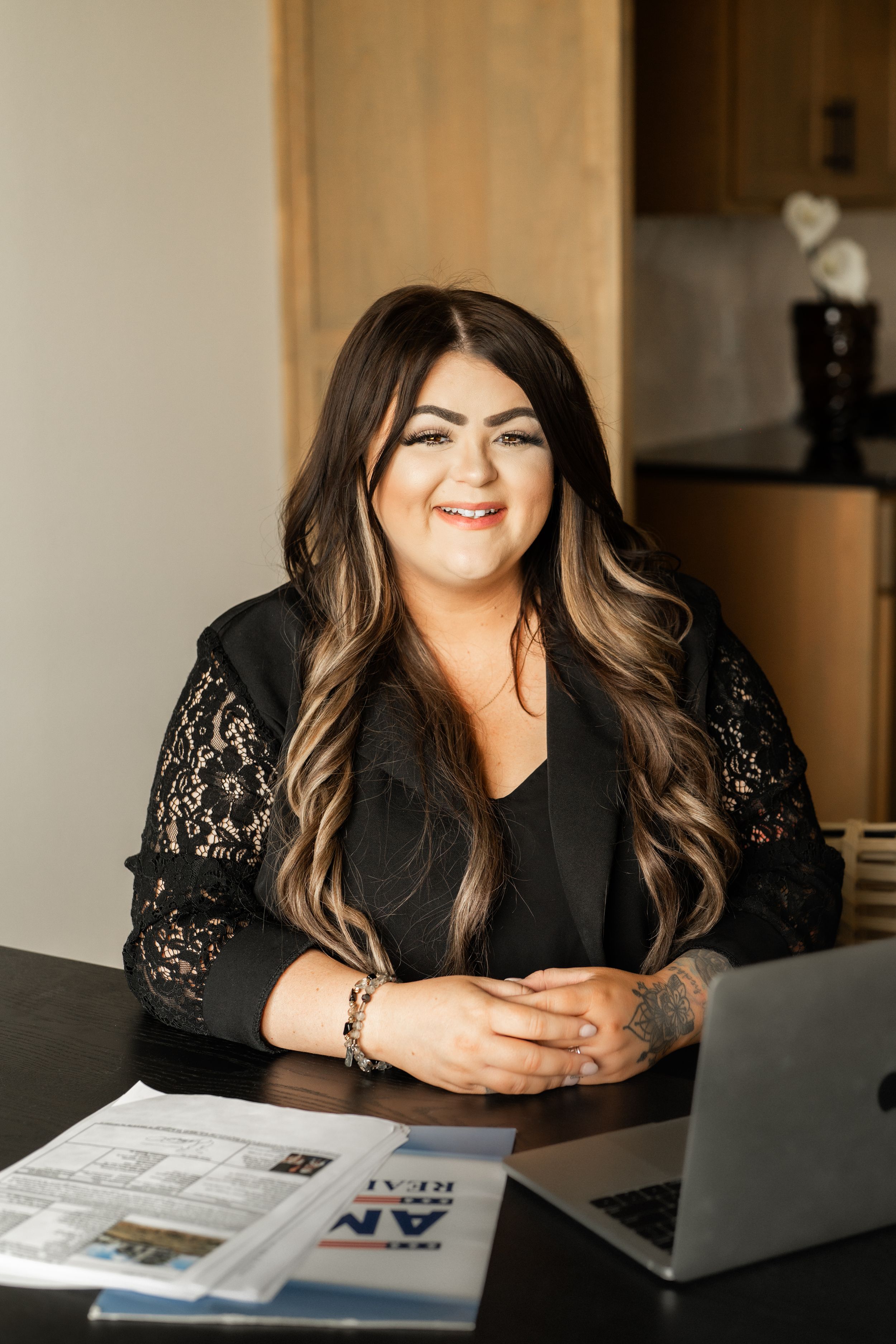 Smiling female real estate agent in black lace top sitting at desk with laptop and property brochures.