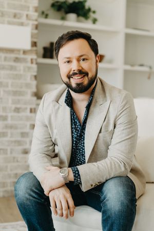 Smiling bearded man in blazer and jeans sits relaxed in modern white interior setting.