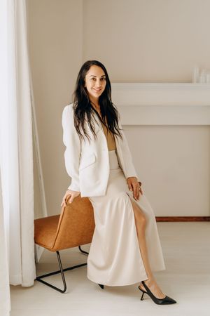 An elegant woman in a white blazer and cream skirt poses on a leather chair in a minimalist space.