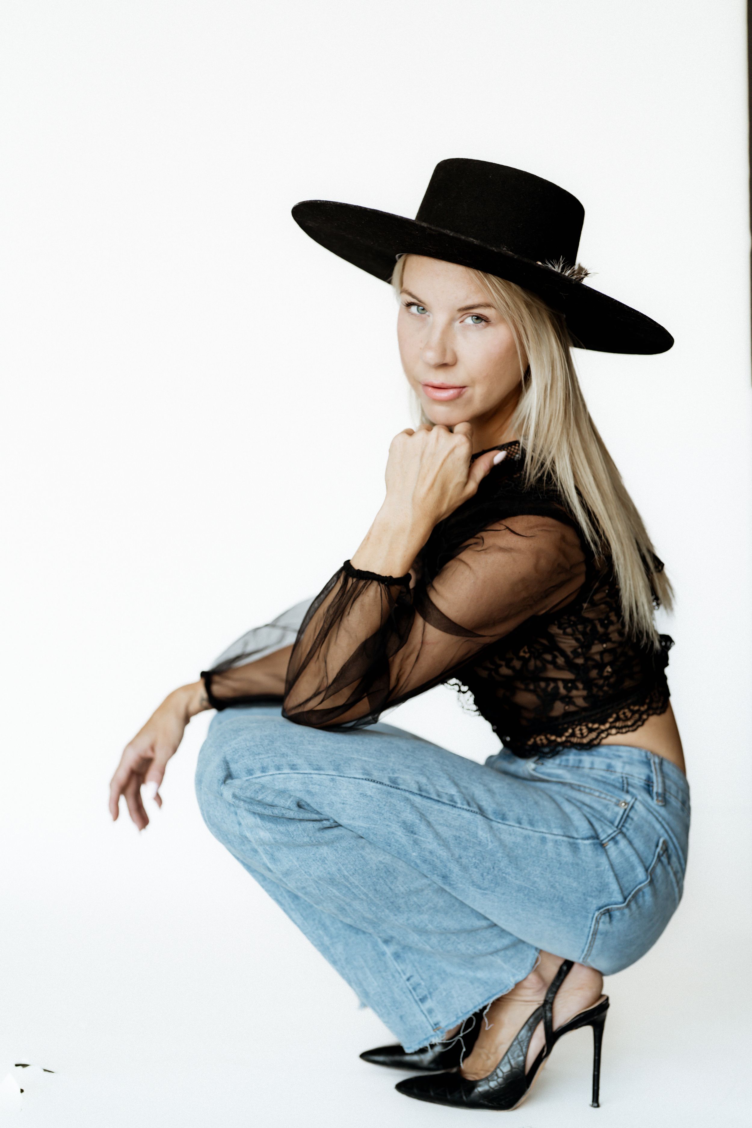 Blonde woman in black wide-brim hat, sheer lace top, blue jeans, and black heels posing on white background.
