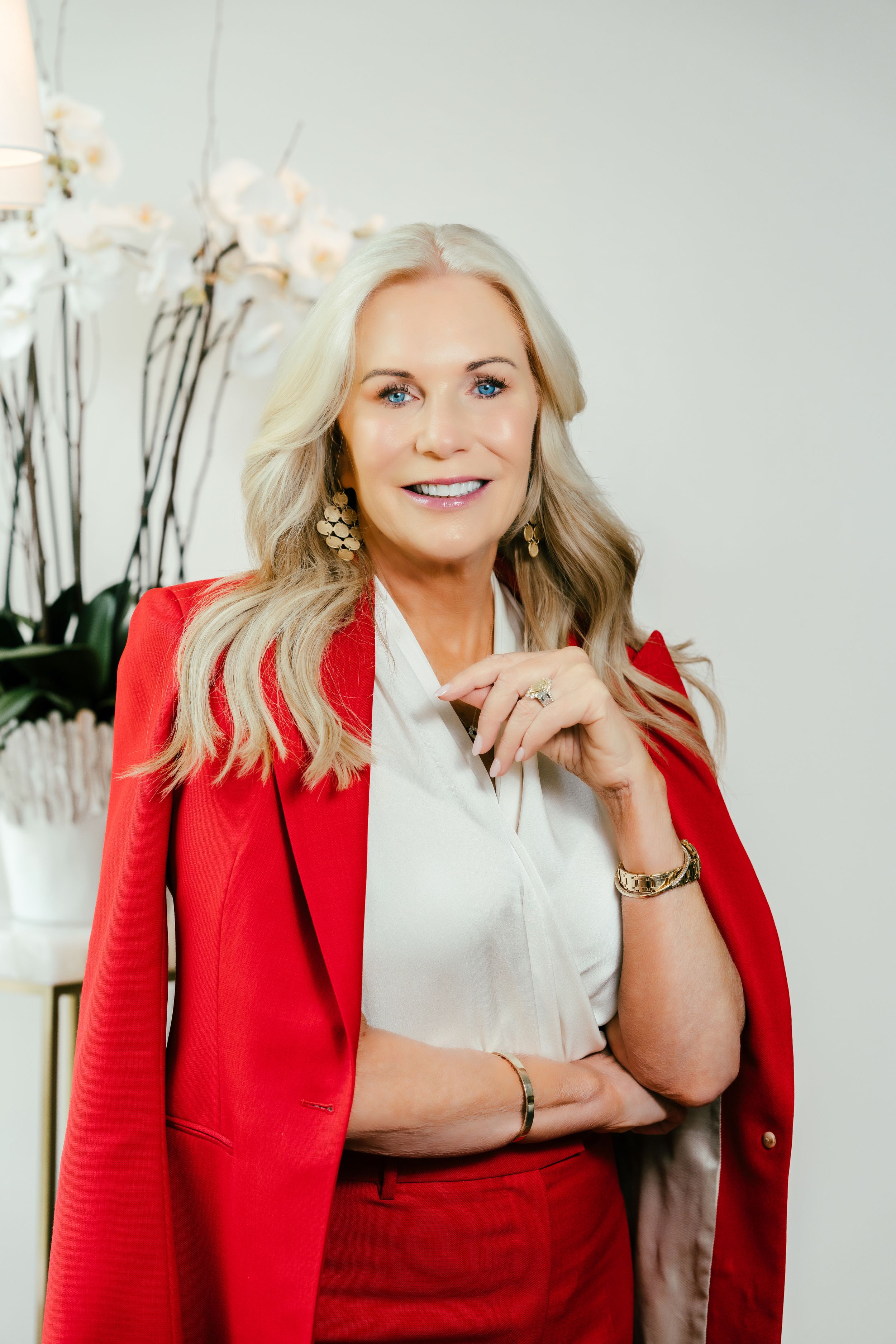 Confident woman in red blazer and pants with white blouse, blonde hair, gold jewelry, smiling indoors.