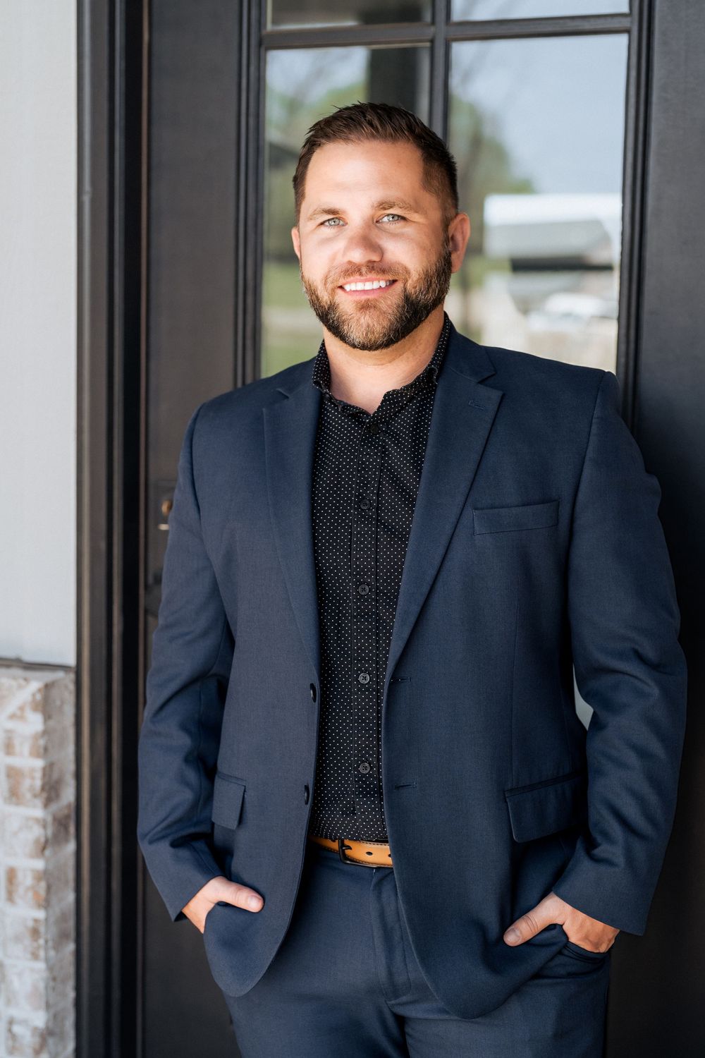 A smiling man in a navy blue suit and black shirt stands confidently with hands in pockets near a dark doorway.