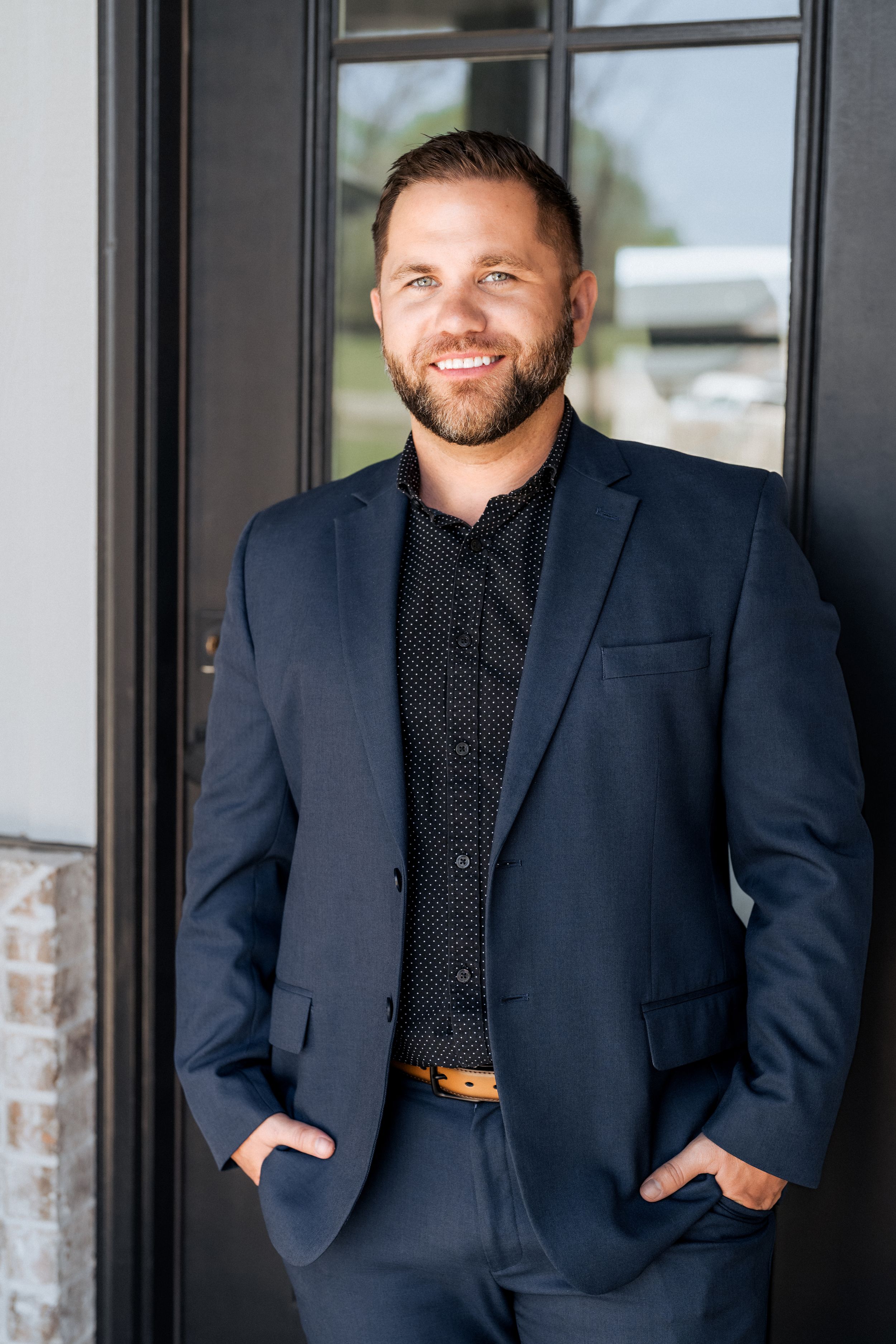 A smiling man in a navy blue suit and black shirt stands confidently with hands in pockets near a dark doorway.