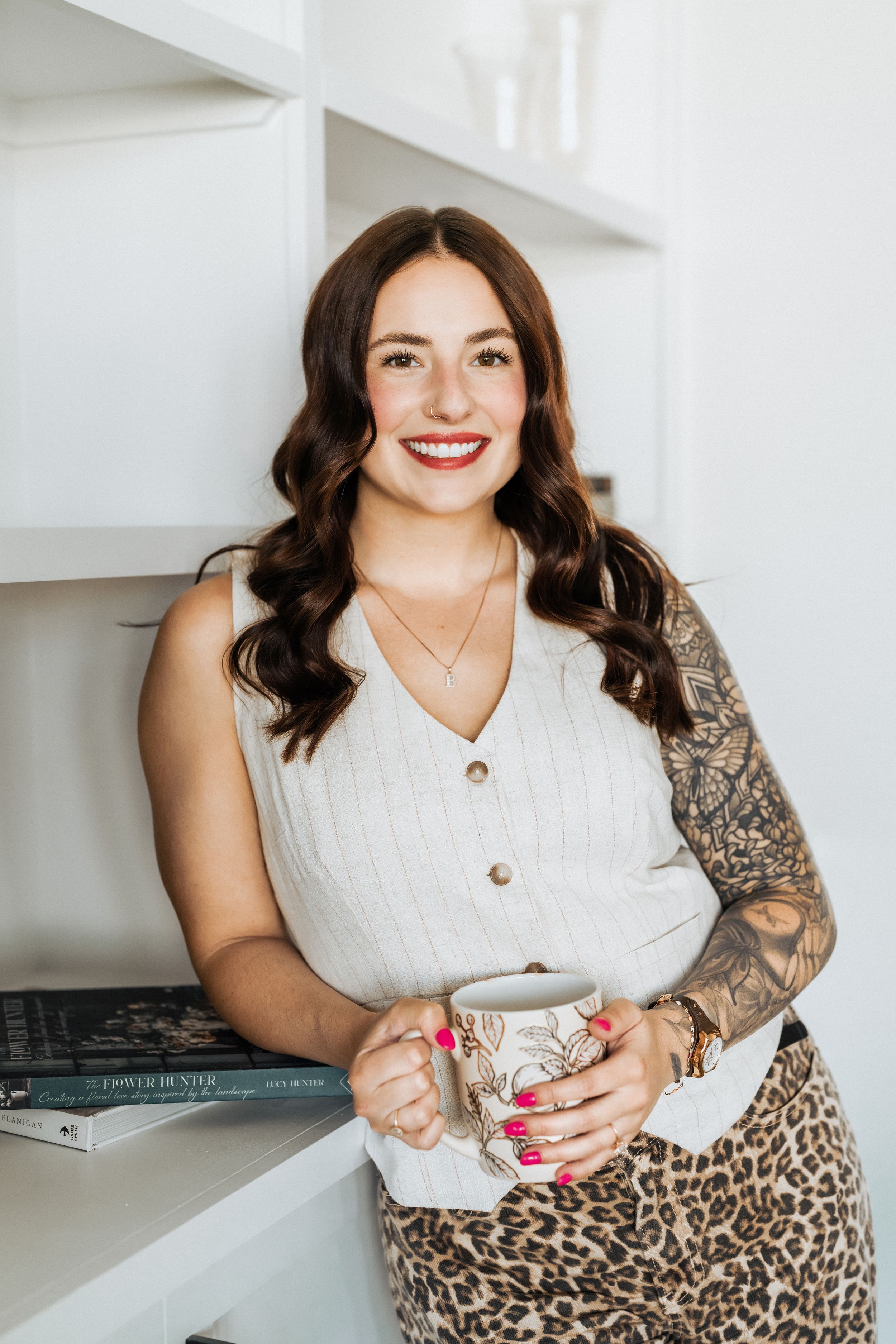 Smiling brunette woman with tattoos holding a coffee mug, wearing a white top and leopard skirt, leaning against white shelves.