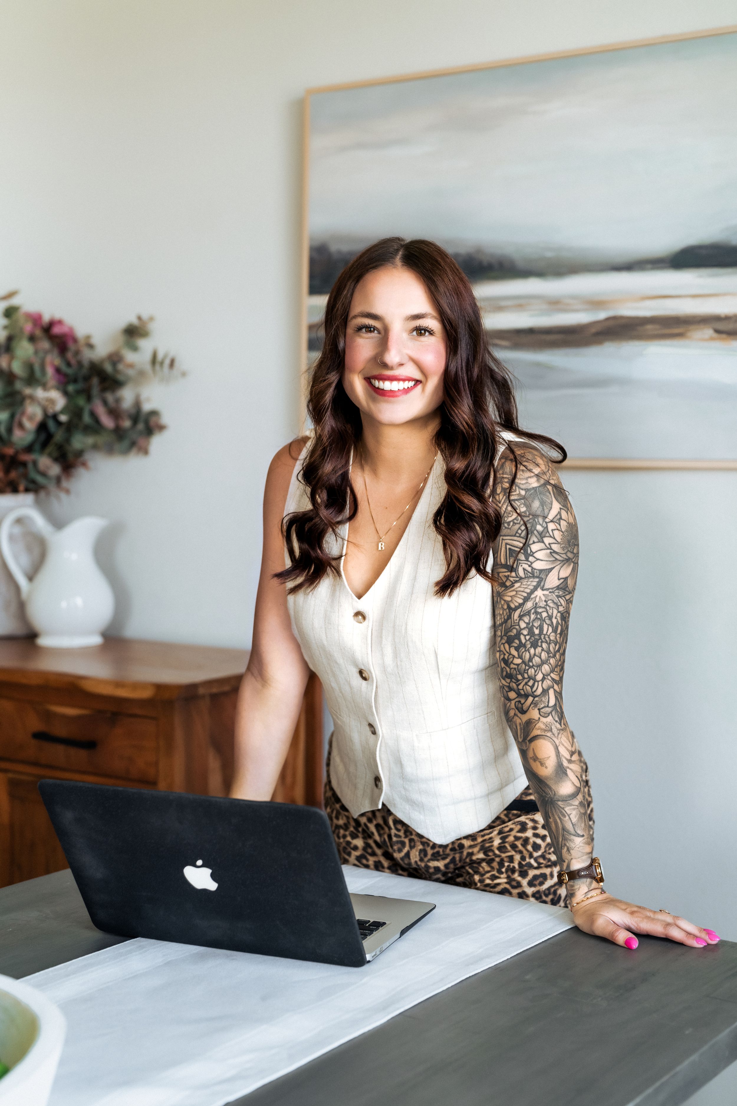 Smiling woman with tattoo sleeve leans on desk with MacBook laptop in bright modern home office.