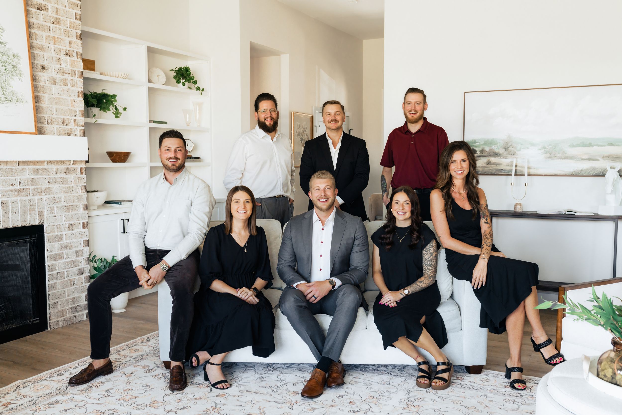 A professional real estate team of eight adults posing together in a modern living room with white built-in shelves.