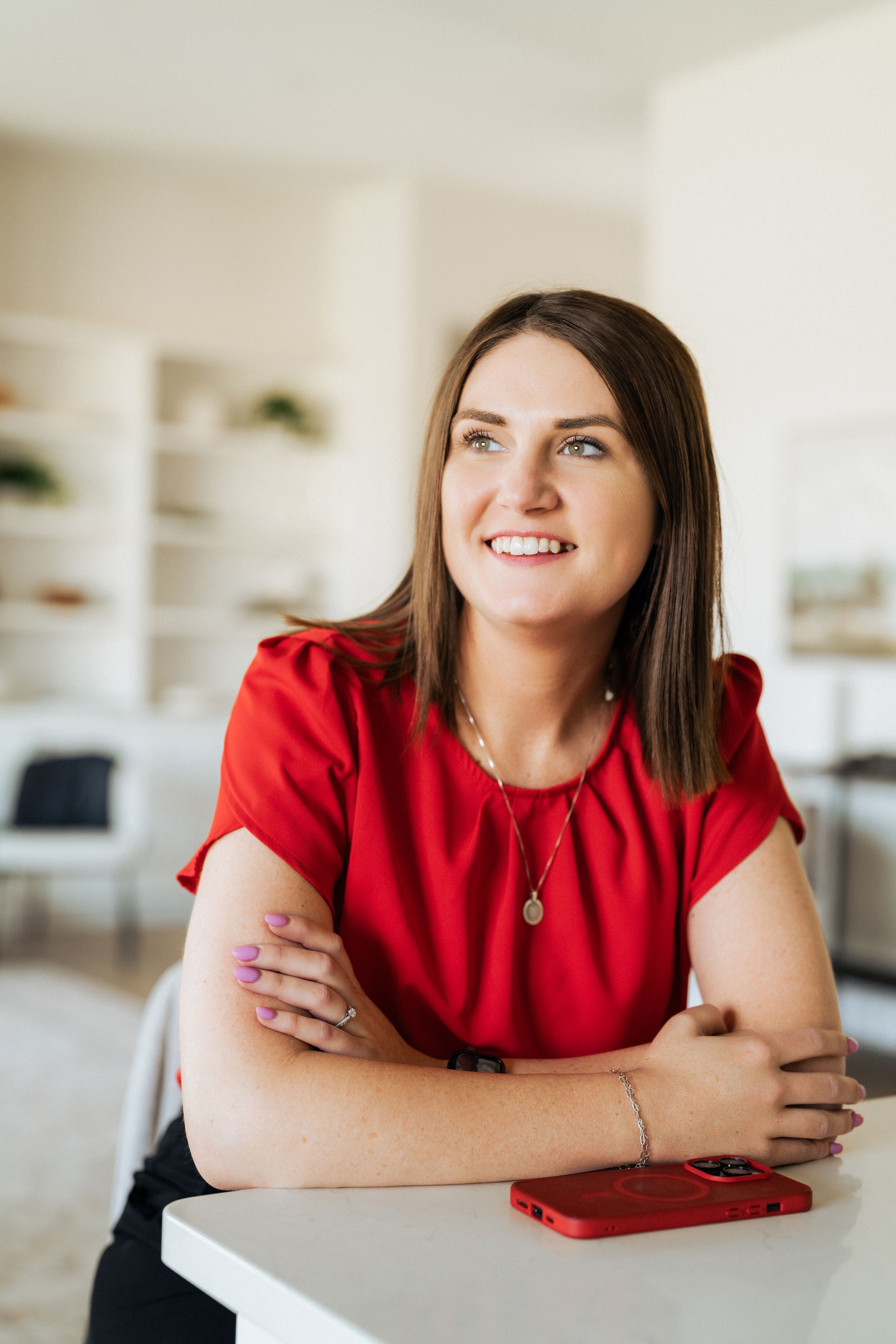 Smiling woman in red blouse sitting at white table with red phone, looking away in bright modern interior.