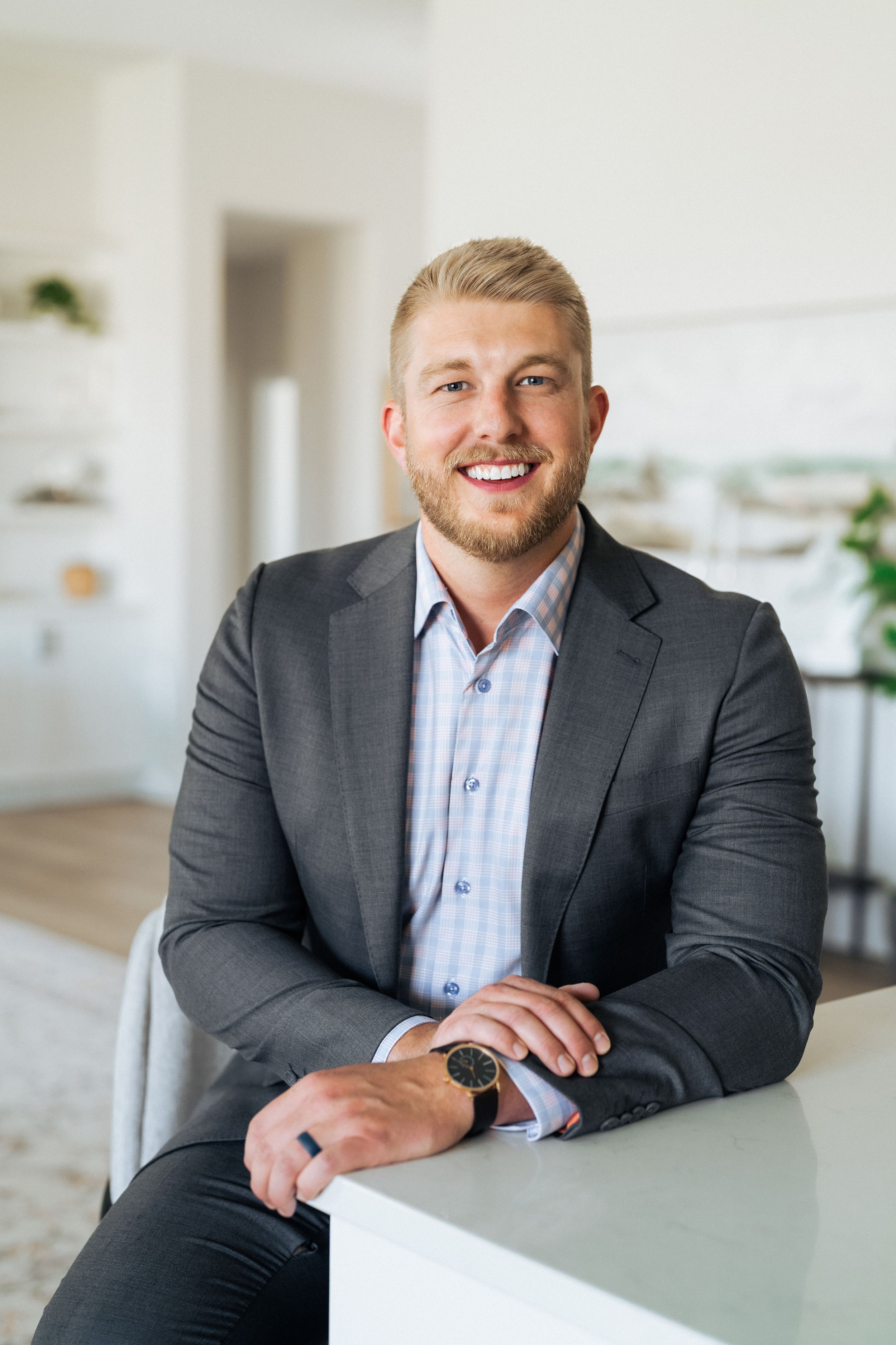 Smiling blonde man in gray blazer and watch leaning on white counter in bright modern interior.