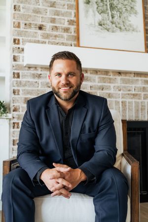 Professional man in navy suit sitting in chair smiling, brick fireplace background, business portrait photo.
