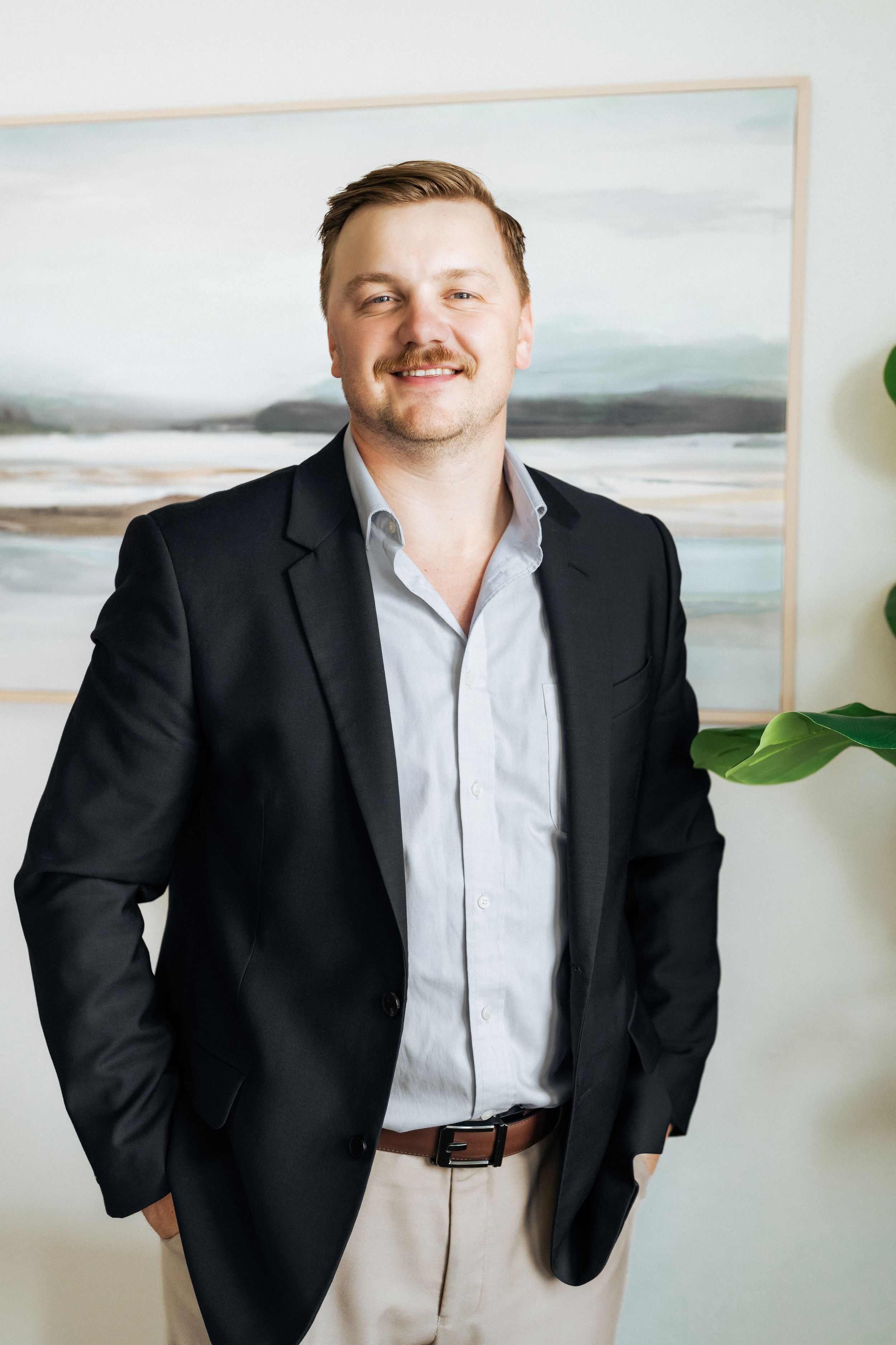 Smiling man with mustache in black blazer and light shirt stands confidently with hands in pockets before coastal artwork.