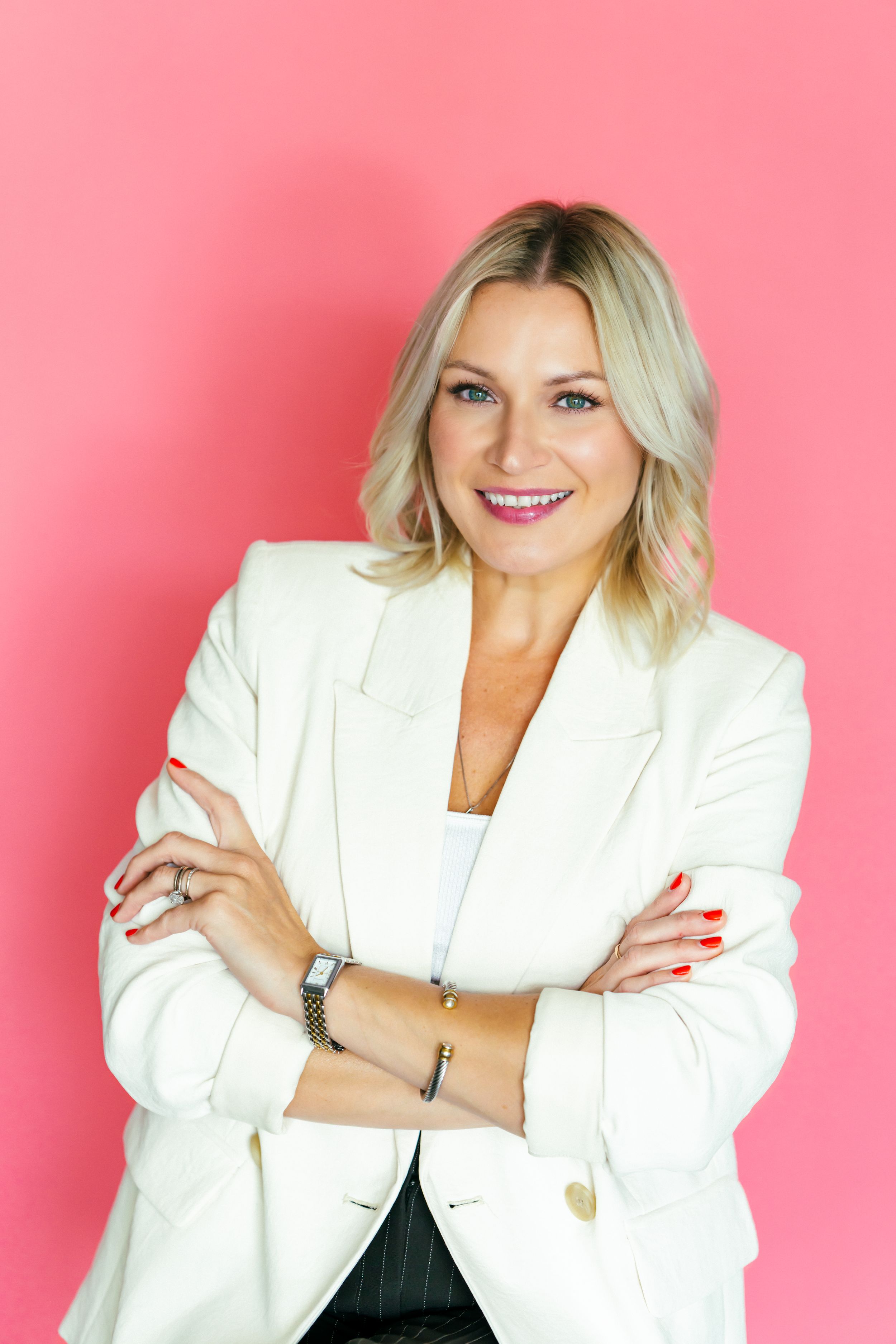 Confident blonde woman in white blazer with arms crossed, smiling against a pink background.