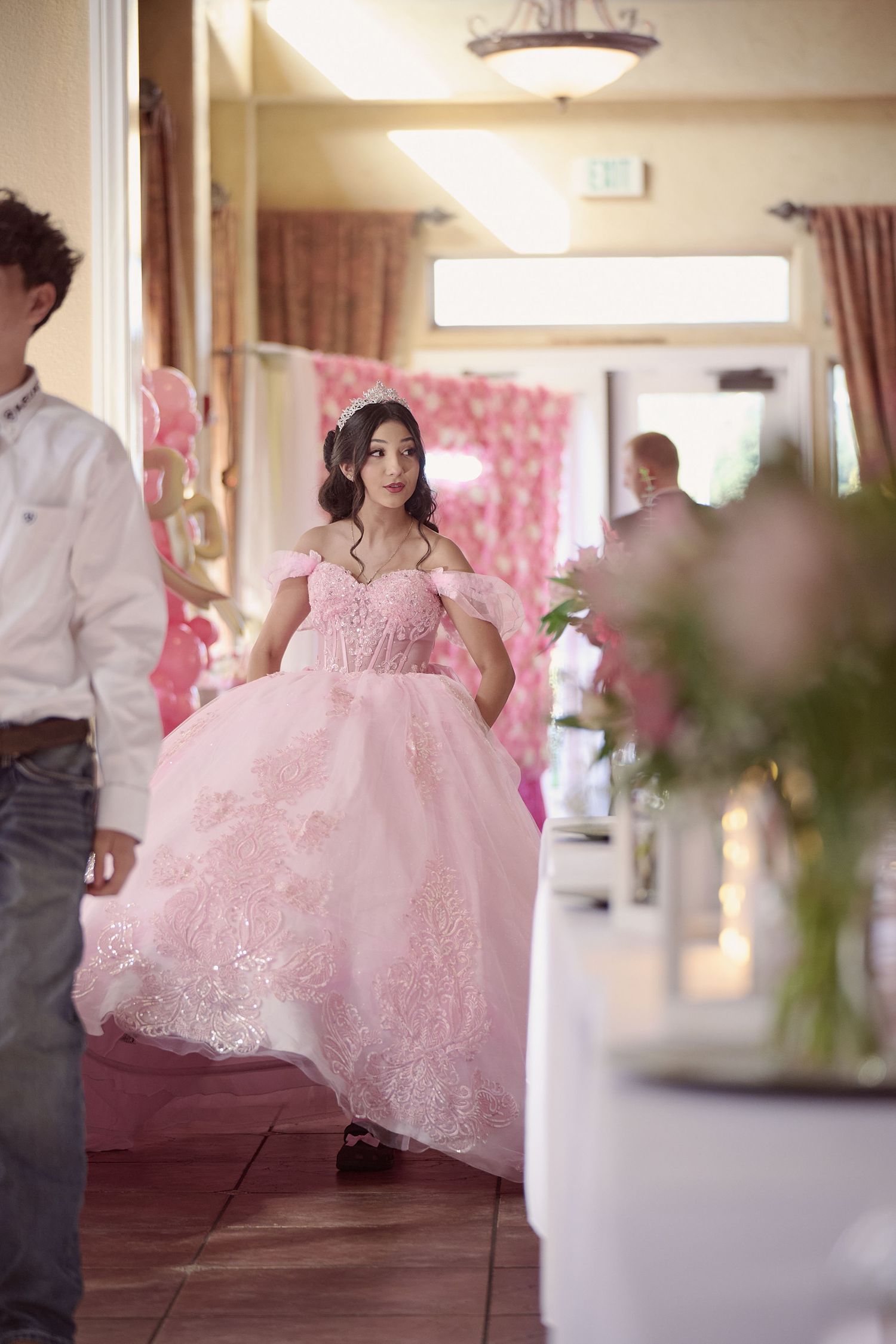 A person wearing a flowing pink lace quinceañera dress twirls in an indoor venue decorated with pink accents.