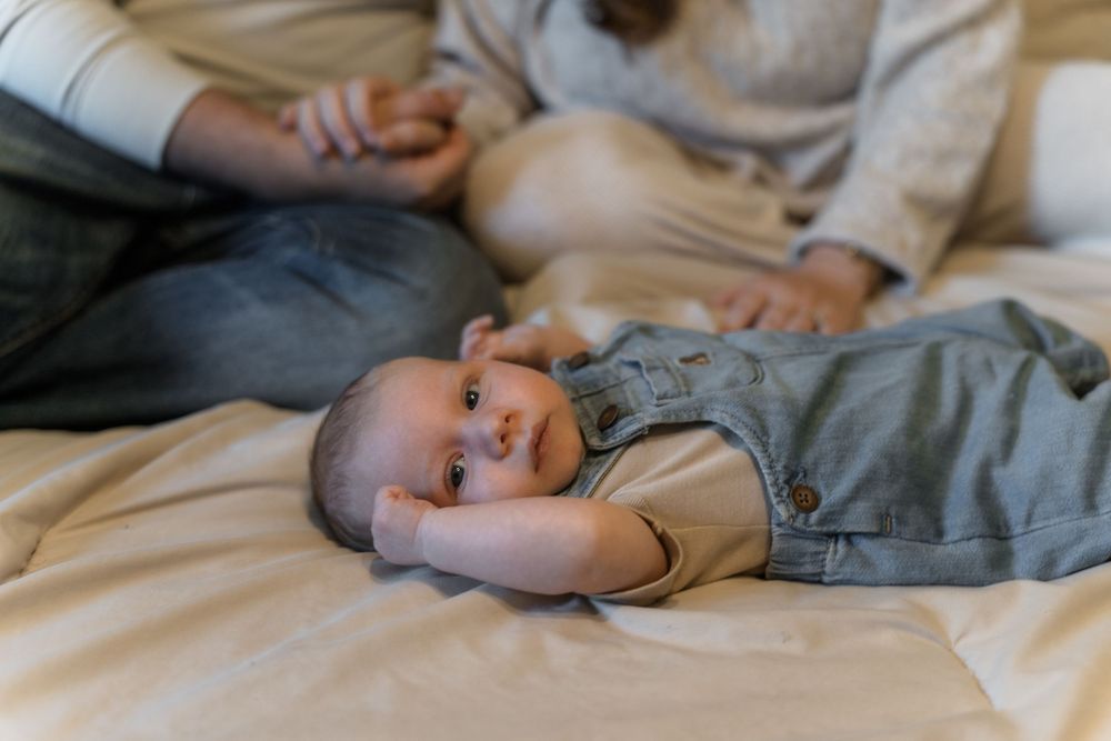 A baby lies on a bed wearing a gray onesie while looking to the side with a curious expression in Macedonia, Ohio in-home newborn photo