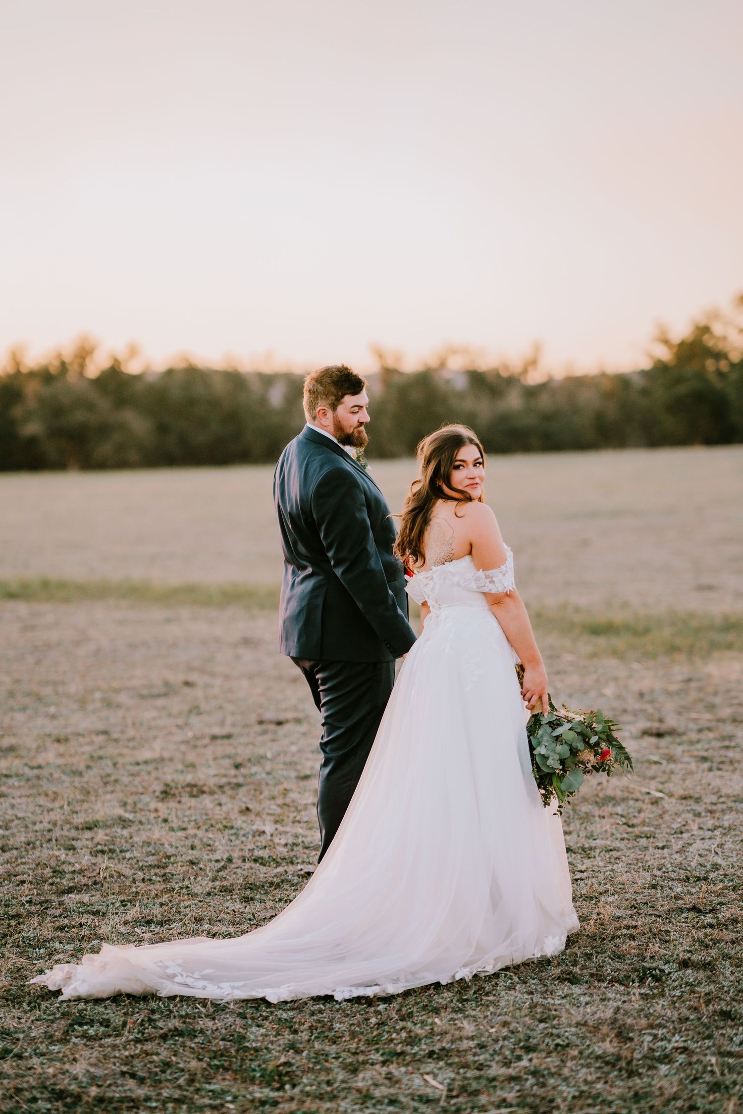 Couple walks hand in hand across rustic field during sunset wedding photoshoot.