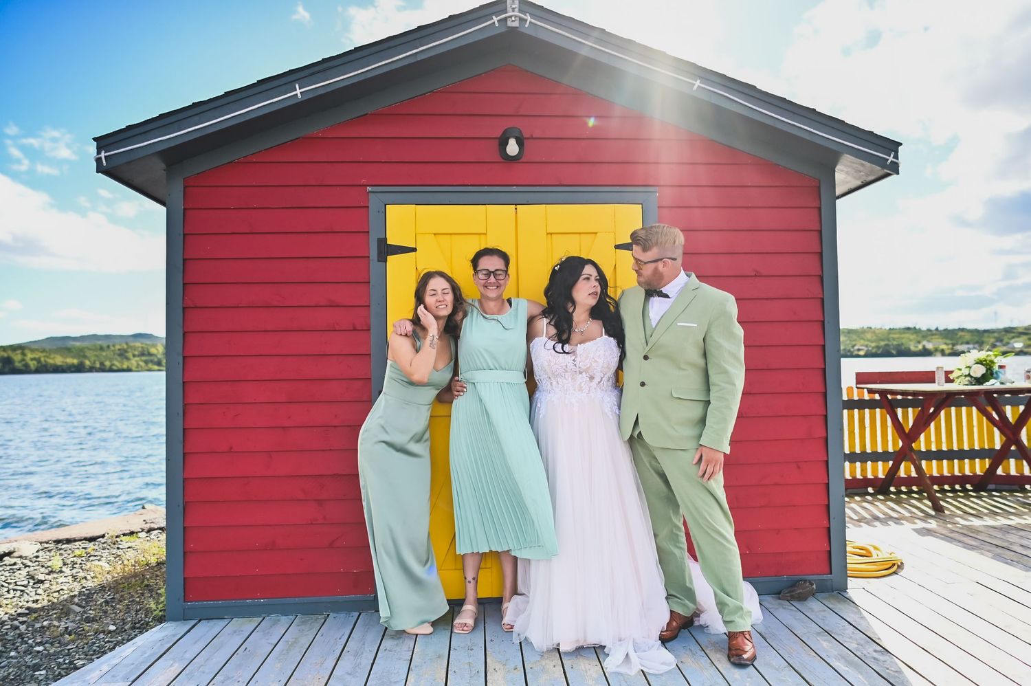 Bridal party in Arnolds Cove in front of a backlit red fishing shed with a yellow door, wearing emerald outfits, bride in a white dress