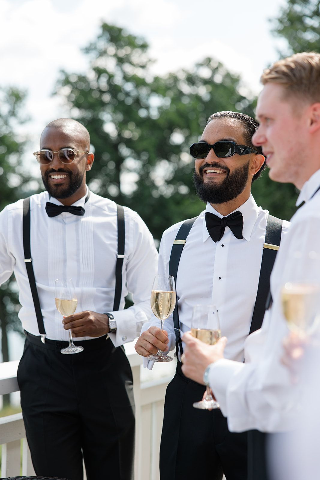 Groomsmen in black tuxedos and bow ties share laughs while holding champagne glasses at an outdoor wedding celebration.