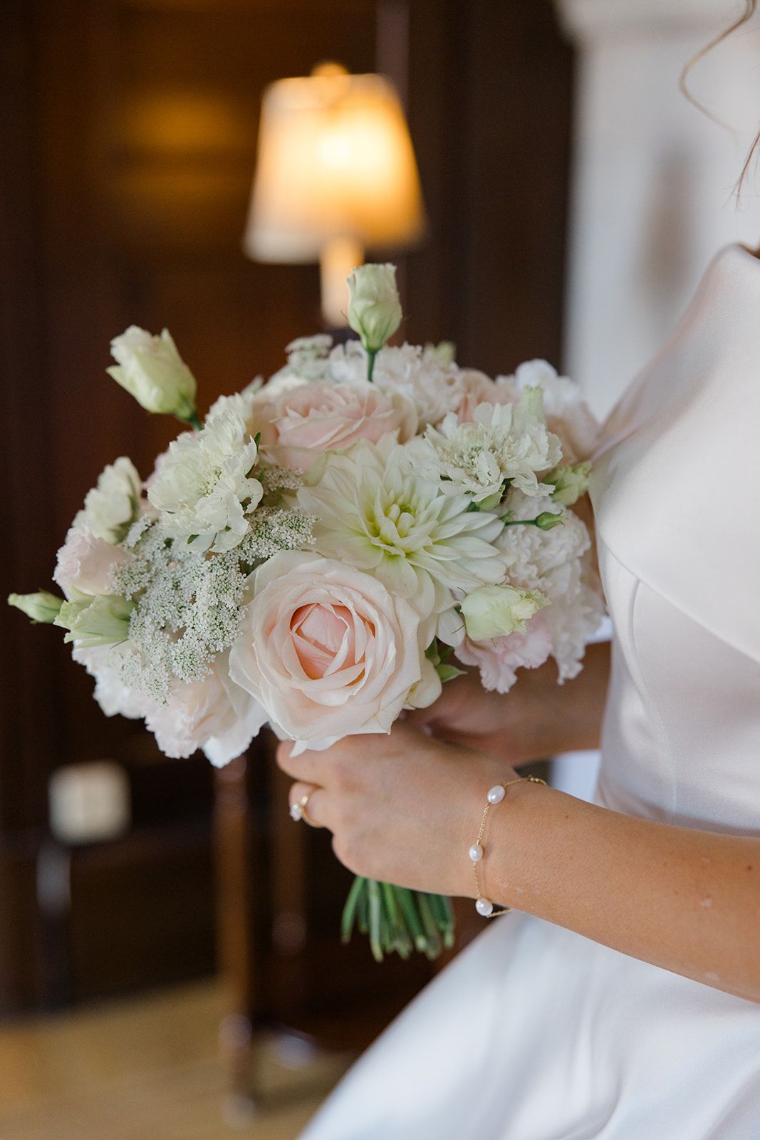 A delicate bridal bouquet of blush pink roses and white carnations held against a white dress with a glowing lamp in background.