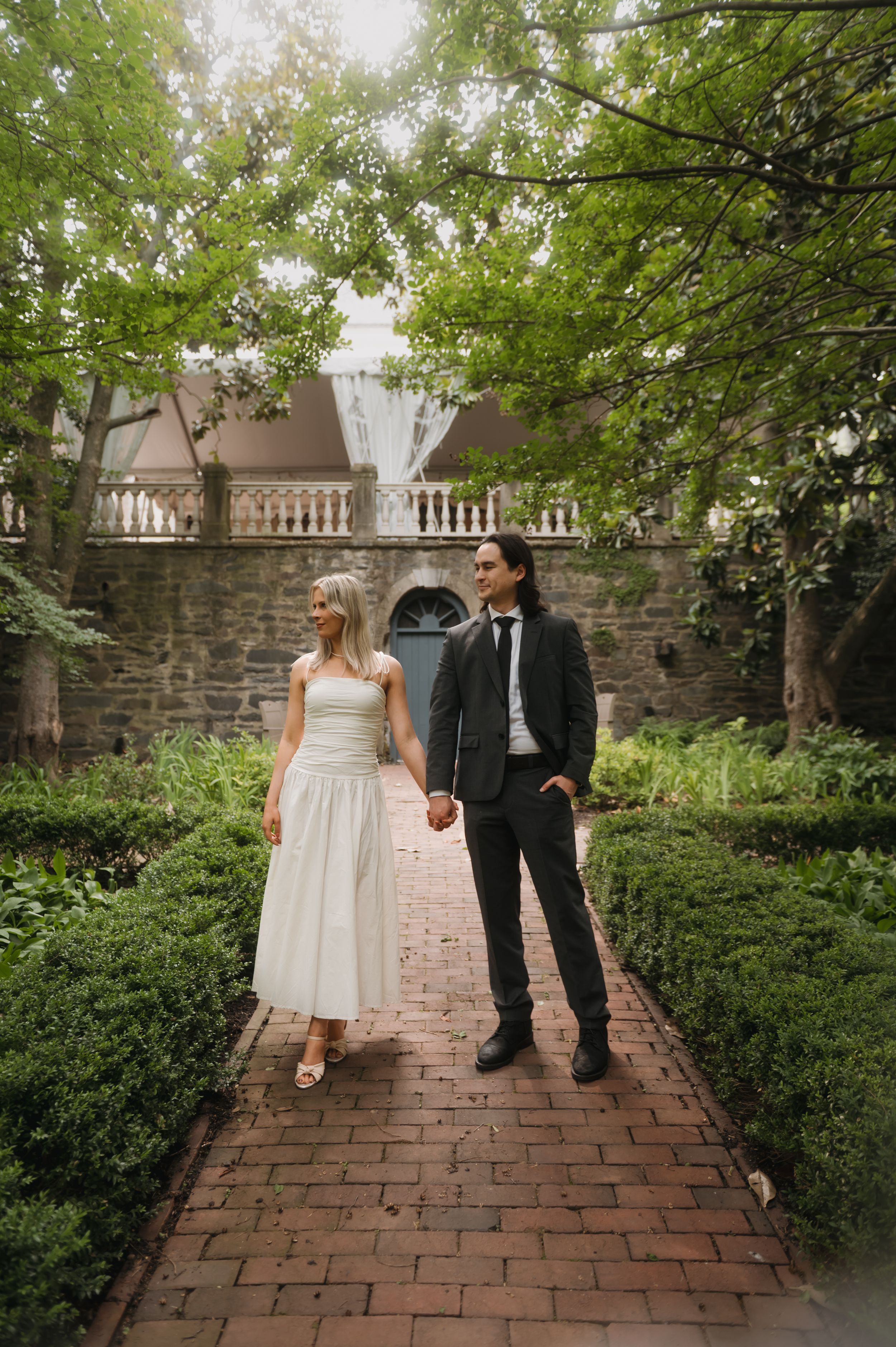 A couple stands on a brick pathway surrounded by lush gardens and stone walls on their special day.