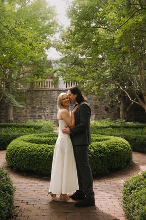 A romantic couple embraces in a formal garden with manicured hedges, brick pathways and stone walls on their wedding day.
