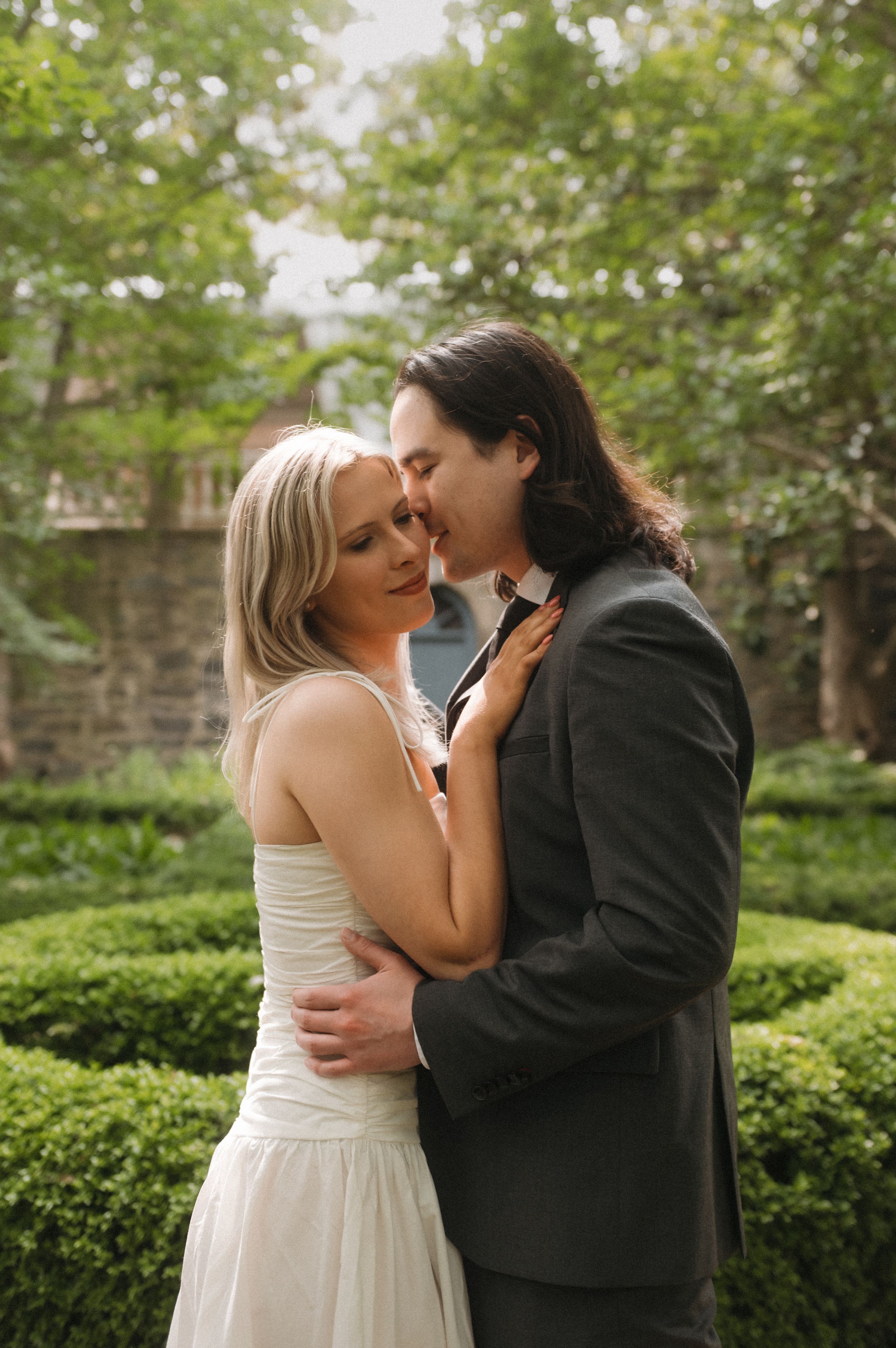A romantic intimate wedding photo showing a couple embracing in a manicured garden with stone walls and greenery.