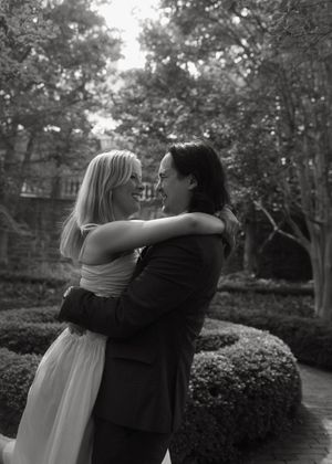 A romantic black and white photograph of an intimate embrace in a garden with manicured shrubs and trees.