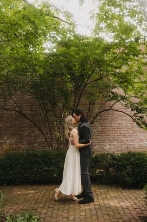 A couple shares a romantic moment in their wedding attire on a brick patio beneath green trees and a historic brick wall.