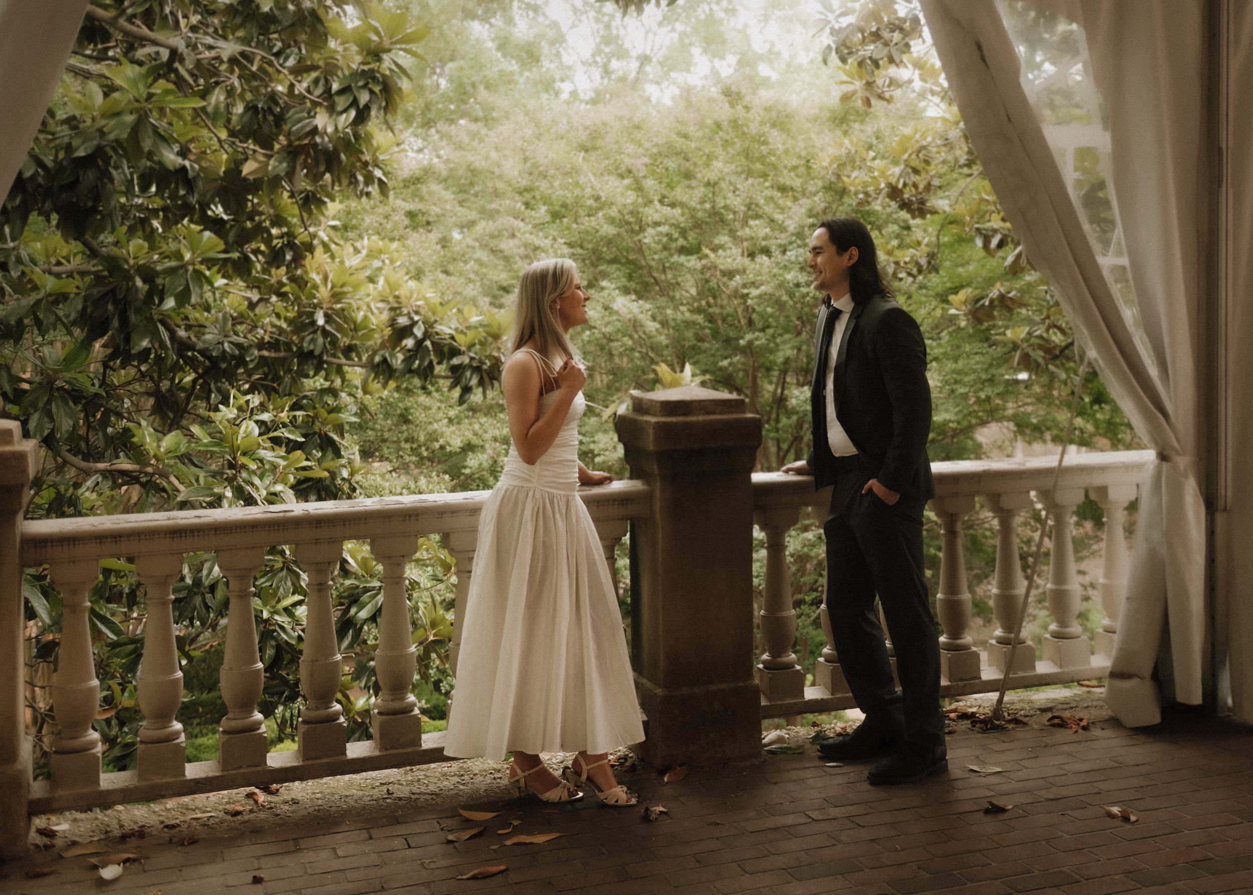 Two people in formal attire stand together on an elegant covered stone balcony surrounded by lush greenery.