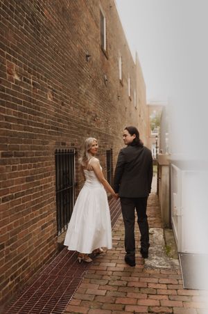 A couple in wedding attire walks together down a narrow brick alleyway beside a historic red brick wall.