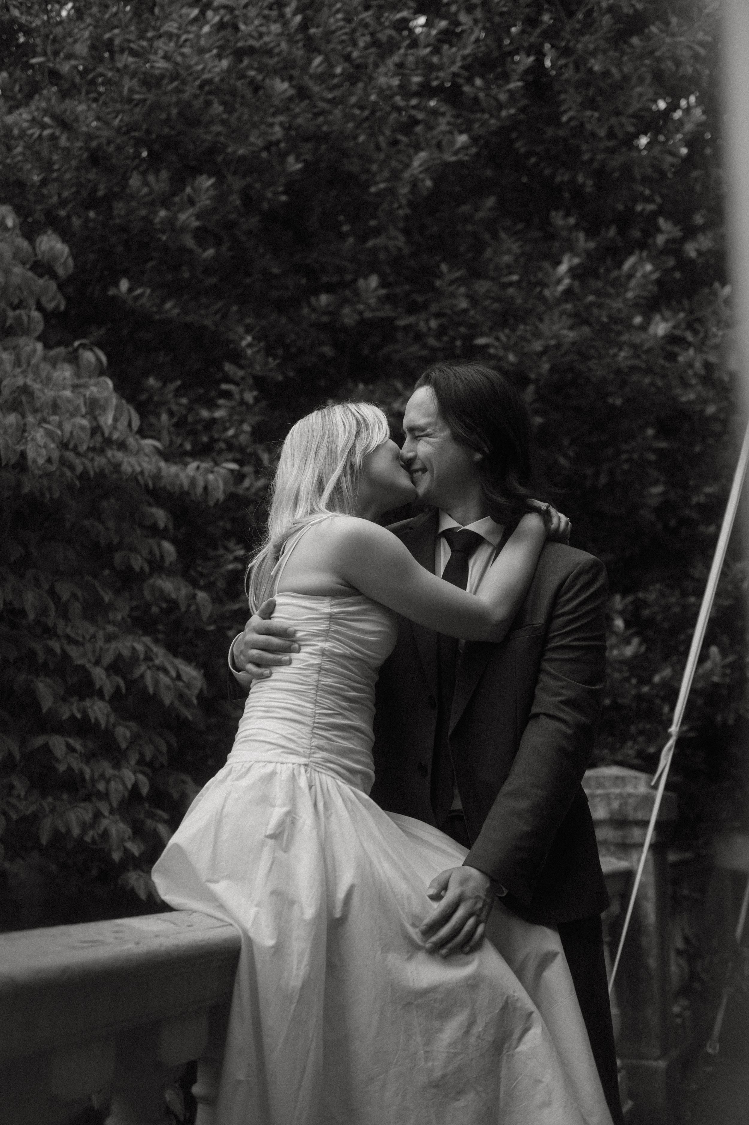 A romantic black and white wedding photograph of a couple embracing on a balcony with greenery in the background.