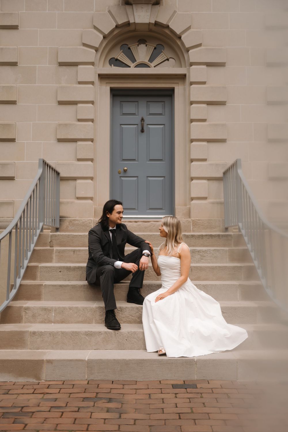 Couple sitting on stone steps in front of an elegant blue door, bride wearing white gown and groom in dark attire.