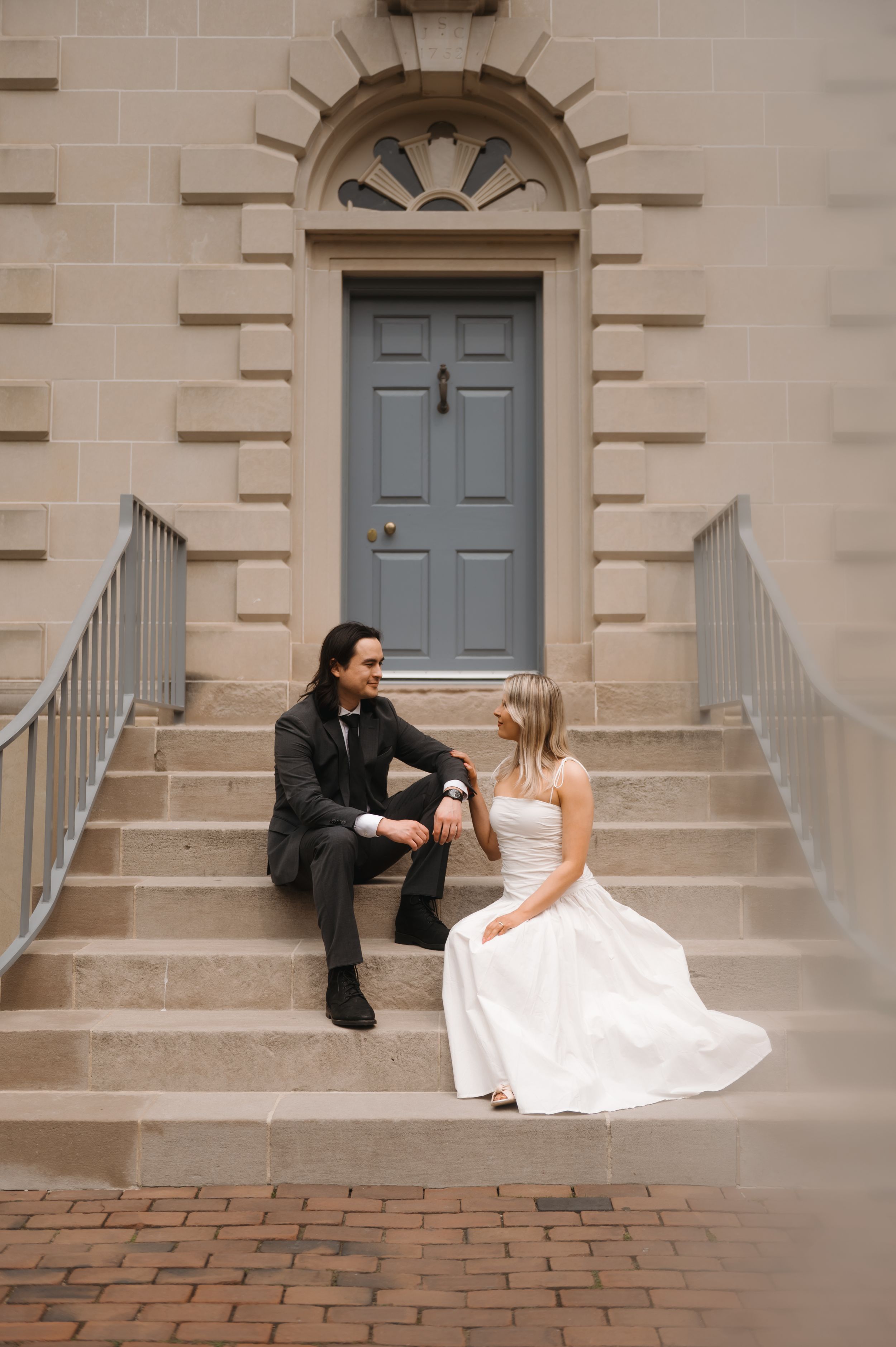 Couple sitting on stone steps in front of an elegant blue door, bride wearing white gown and groom in dark attire.