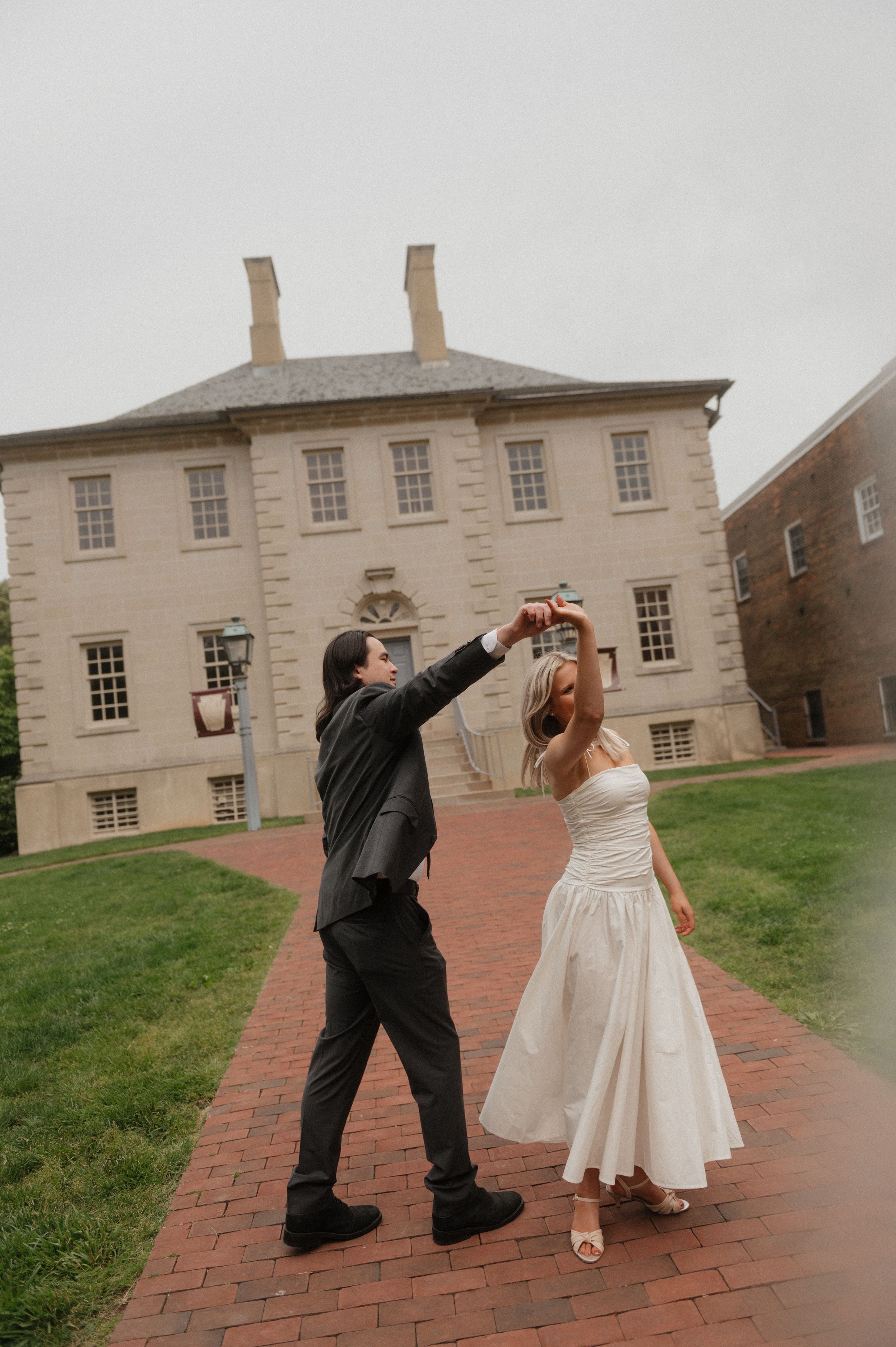 A couple dance together on a brick pathway in front of a historic Georgian-style colonial building.