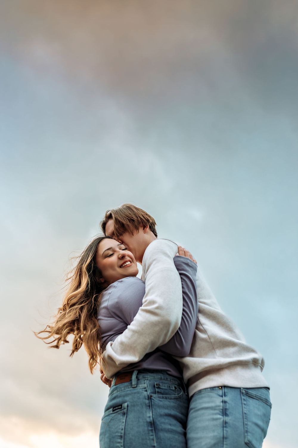 Intimate moment shared between couple in casual attire against soft cloudy sky.