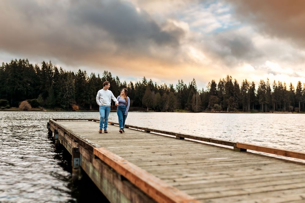 Long wooden pier stretches over calm lake waters at sunset with pine forest shoreline.