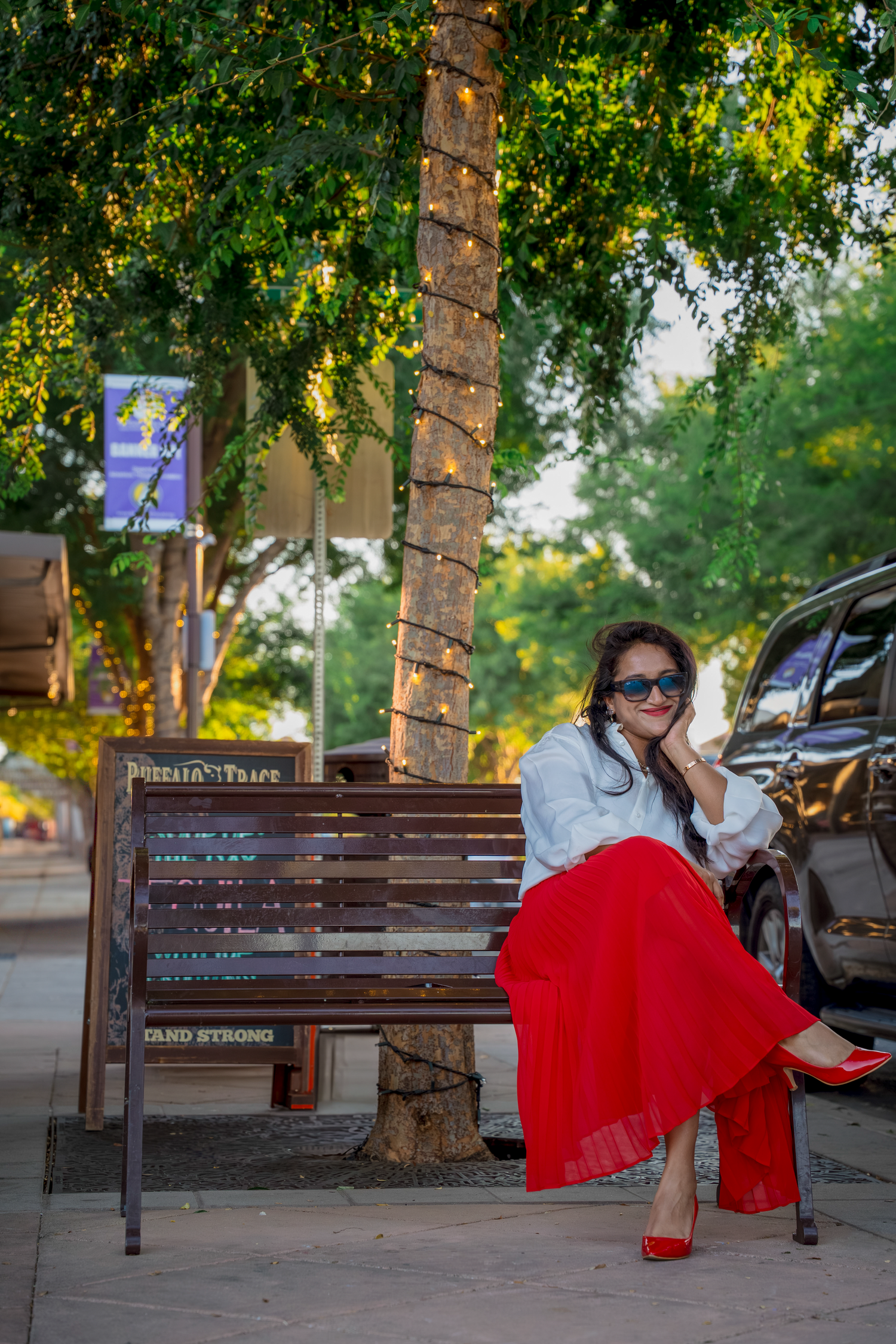 Person in a flowing red dress poses playfully on a wooden bench under trees in an urban setting.