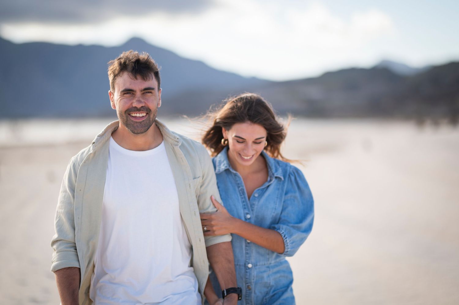 A couple walks hand in hand along a sandy beach trail with mountains and blue sky in the background.