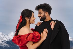 A romantic couple embraces outdoors, with the woman wearing a flowing red dress contrasting against a cloudy sky.