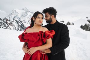 A couple in formal attire embraces against a snowy mountain backdrop with the woman wearing a vibrant red gown.