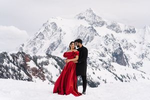 A romantic couple poses in the snow-covered mountains with the bride wearing a flowing red dress against white peaks.