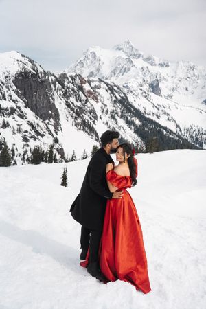 A couple in formal winter attire embrace against a backdrop of snow-covered mountains in a romantic winter photoshoot.