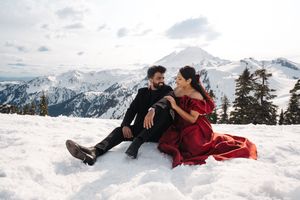 Two people lay close together in the snow wearing winter coats against a scenic mountain backdrop.