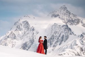 A couple in red winter coats takes photos while enjoying scenic snowy mountain peaks in the background.