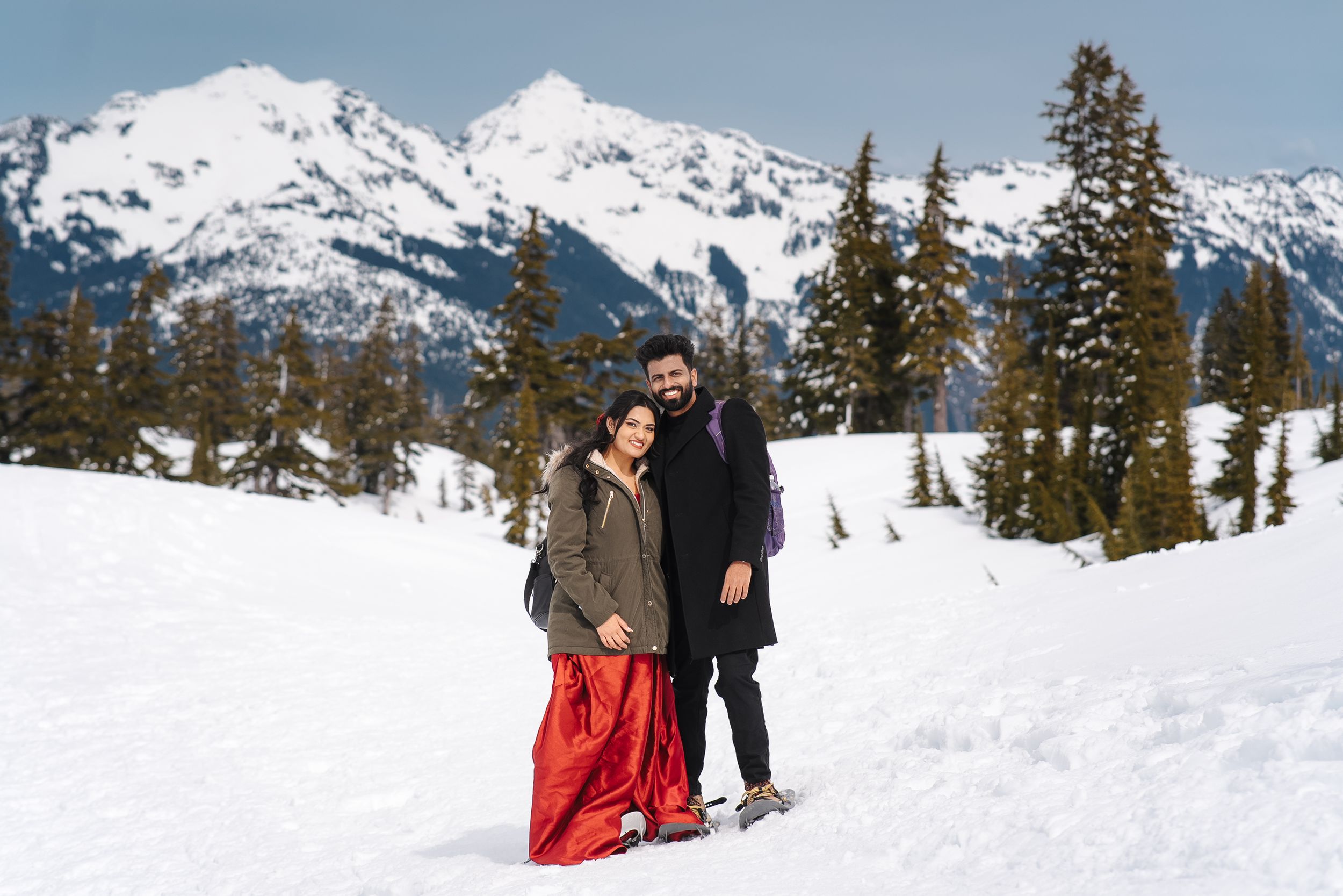 Two people stand together in snowy mountain landscape with evergreen trees and snow-capped peaks in background.
